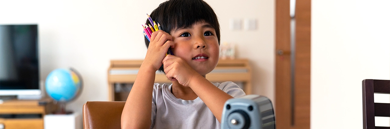 Criança pequena sentada em uma mesa dentro de casa segurando um maço de lápis de cor ao lado do rosto.