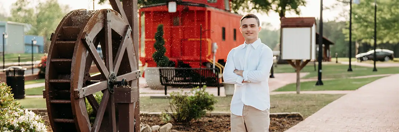 Un estudiante de último año de secundaria posa al aire libre junto a una noria de madera, con un vagón de tren rojo al fondo, durante una sesión de fotos de graduación.