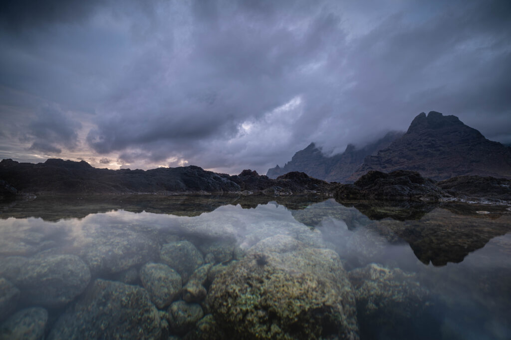 A dramatic seascape with rugged rocks, misty mountains, and a reflective tide pool, captured in stunning detail using Tamron lenses for Sony A7R series cameras.