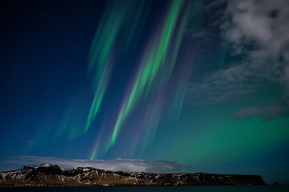 Vibrant green and purple aurora borealis streak across a starry night sky above a snow-dusted mountain range and coastal village in Iceland, with clouds partially illuminated by the aurora’s glow.