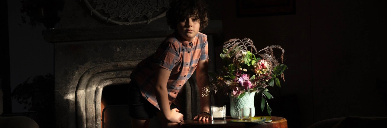 A young girl with curly hair wearing a plaid shirt leans on a table beside a flower arrangement in a dimly lit room, with a beam of light highlighting her face and the bouquet.