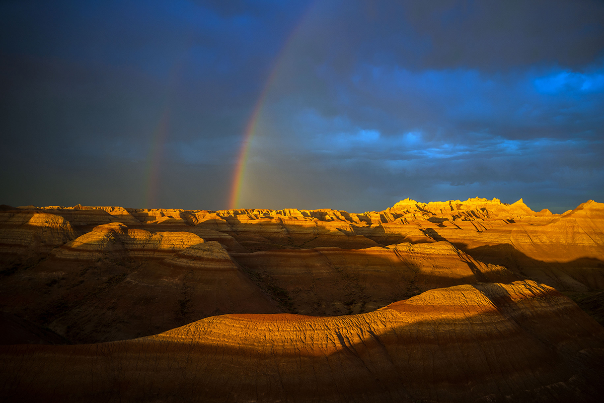 A dramatic desert landscape with layered rock formations, illuminated by sunlight and featuring a double rainbow against a dark sky.