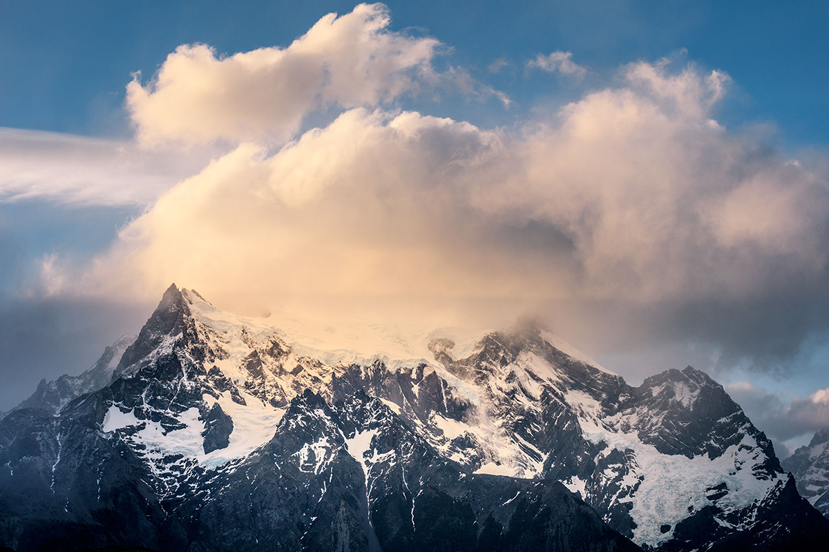 A snow-covered mountain peak under a soft, golden cloud against a blue sky.