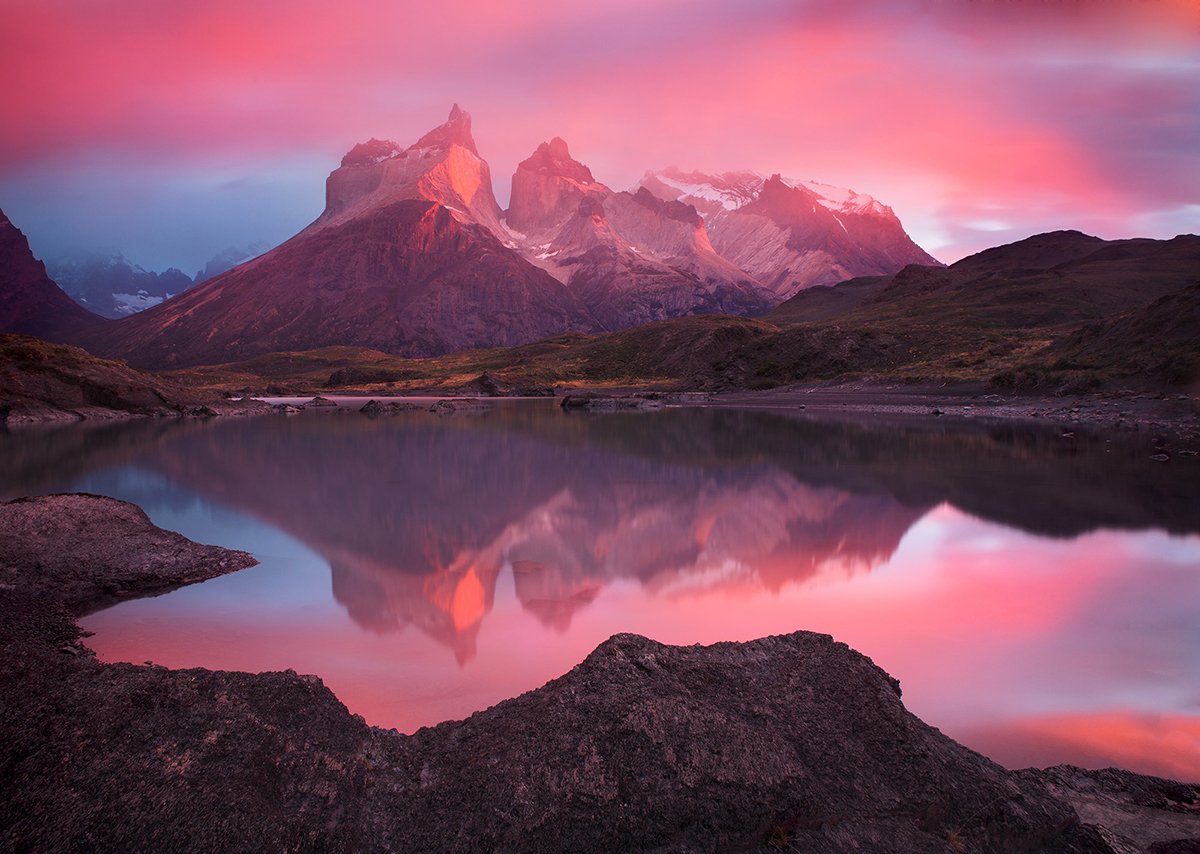 A serene mountain landscape at sunrise or sunset, with pink and purple skies reflecting off a calm lake, surrounded by rugged terrain.