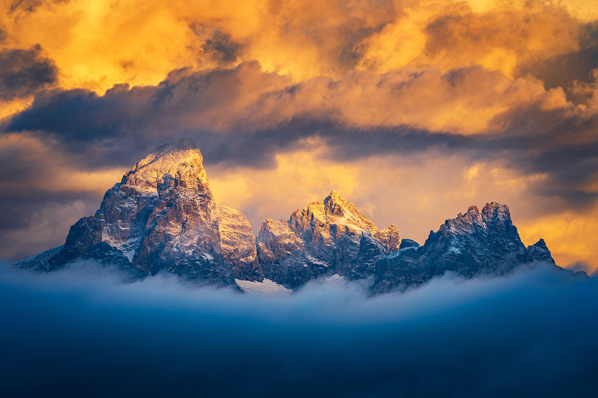 A dramatic mountain peak illuminated by golden light, partially veiled in low clouds with a fiery, orange sky overhead.