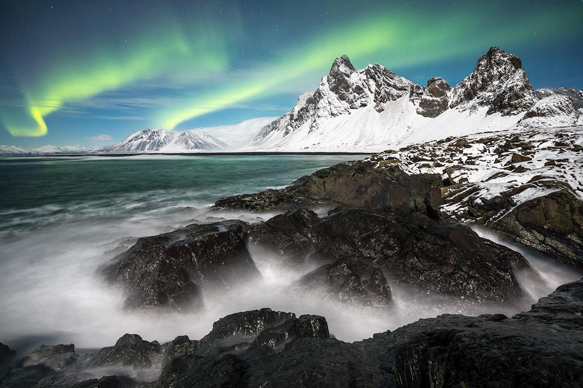 A snowy coastal landscape with rugged black rocks, calm green sea, and vibrant northern lights illuminating the sky.