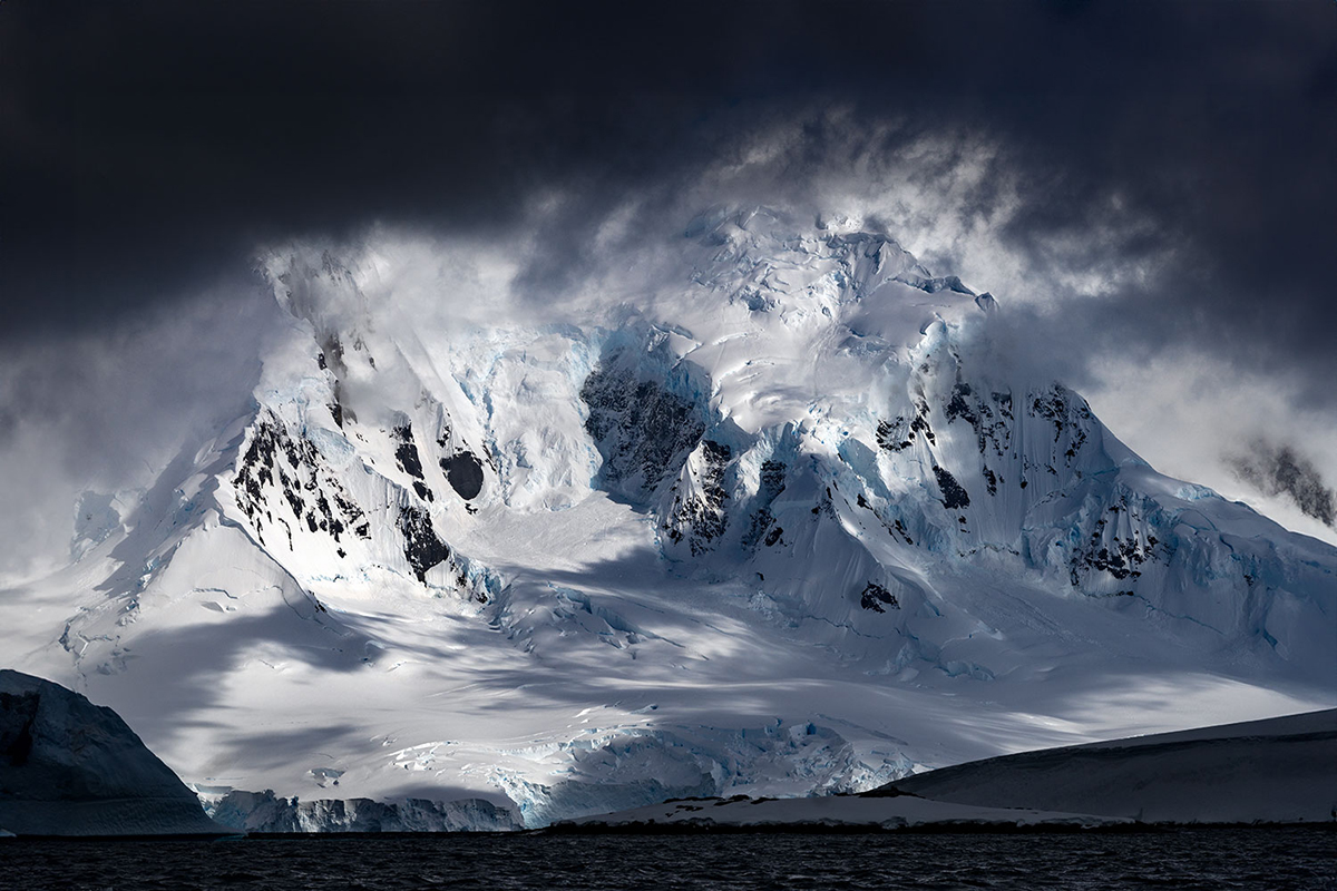 A dramatic snow-covered mountain under dark, stormy clouds with patches of light highlighting icy details.