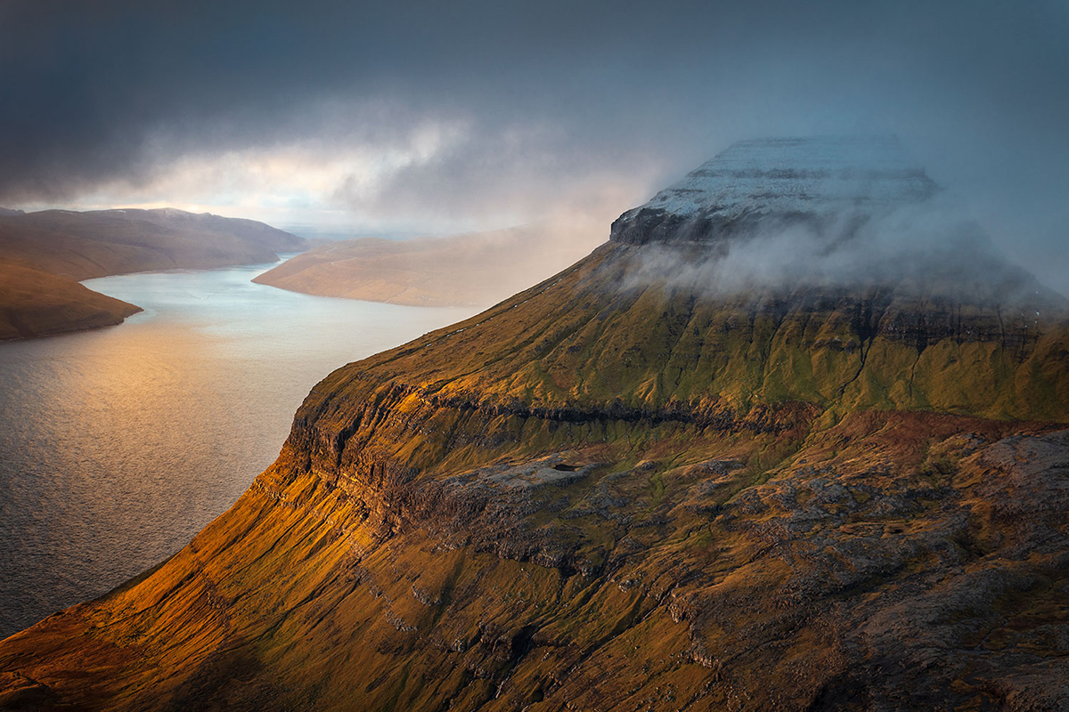 A mist-covered mountain ridge illuminated by sunlight, overlooking a serene body of water with distant hills under a cloudy sky.