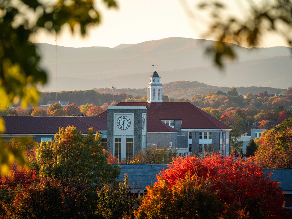 A university building with a clock tower in the foreground, surrounded by vibrant autumn trees in shades of red, orange, and green and against softly lit mountains.