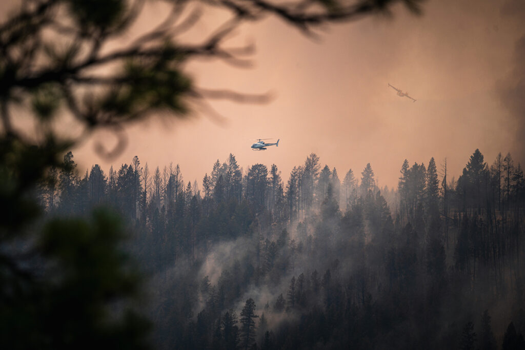 A helicopter and plane fly over a forest engulfed in smoke from a wildfire, with trees silhouetted against a hazy, orange sky.