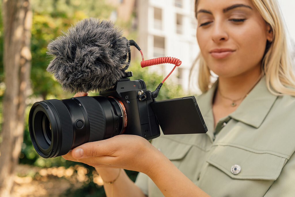 A woman holding a professional camera with a lens and basic videography equipment attached prepares to record outside. 