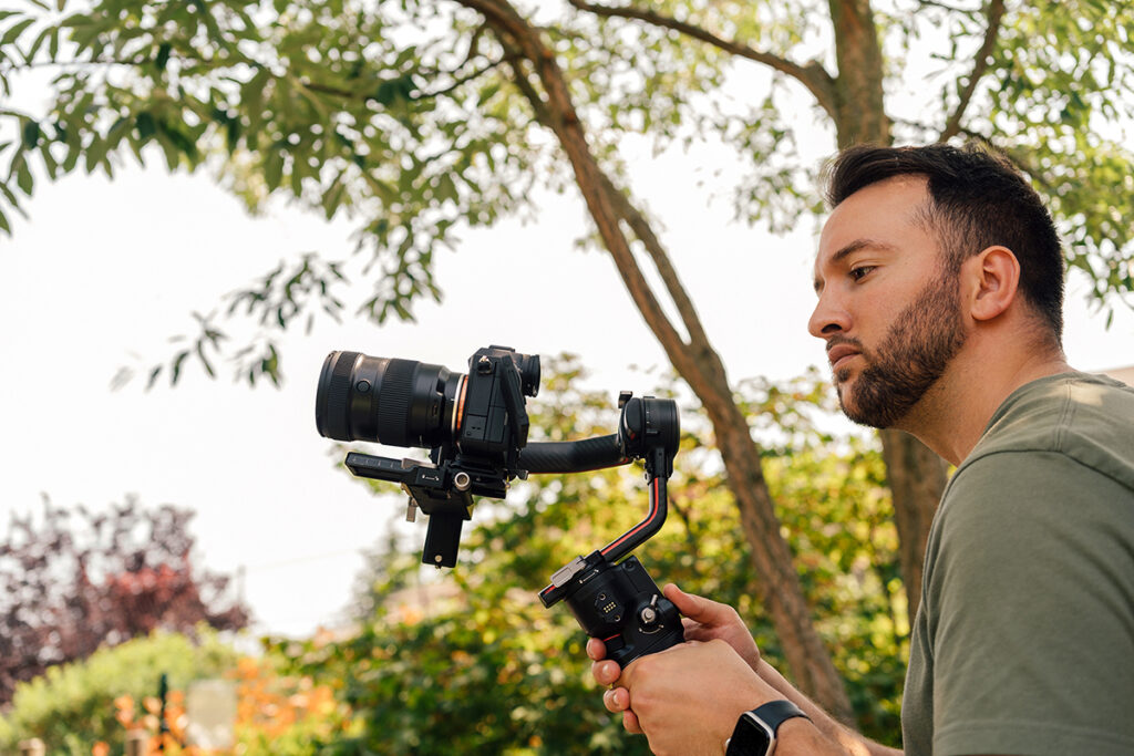 A man holing a camera mounted on a stabilizer gimble and equipped with essential basic videography equipment records video outdoors.