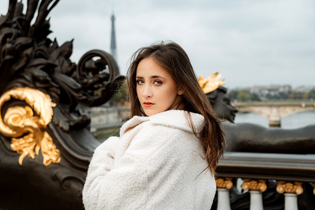 A woman in a white coat stands on an ornate dark brown bridge with gold accents looking over her shoulder to face the camera.