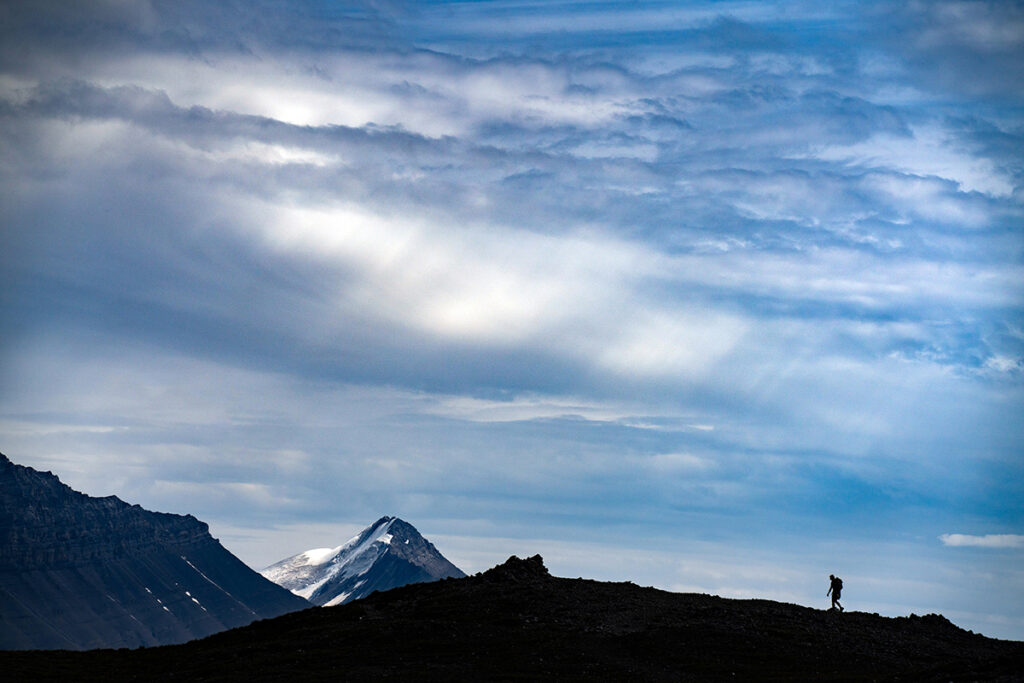 A lone hiker walks along a ridgeline with an expansive sky filled with clouds and a snowcapped peak in the background.