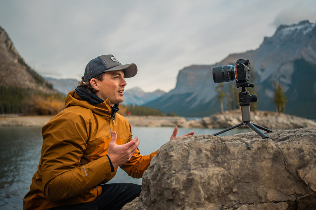 A man sitting outdoors in front of a lake expressively gestures to a camera mounted on a small tripod pointed at him.