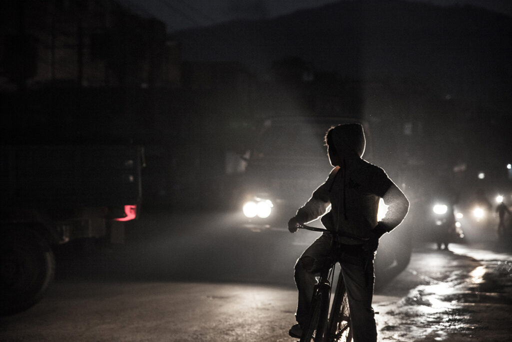 A child on a bike wearing a hooded sweatshirt is silhouetted by the headlights of an approaching vehicle on a dimly lit street. 