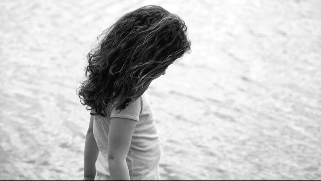 A child with long hair is photographed in black and white while looking downward thoughtfully in front of a calm body of water.