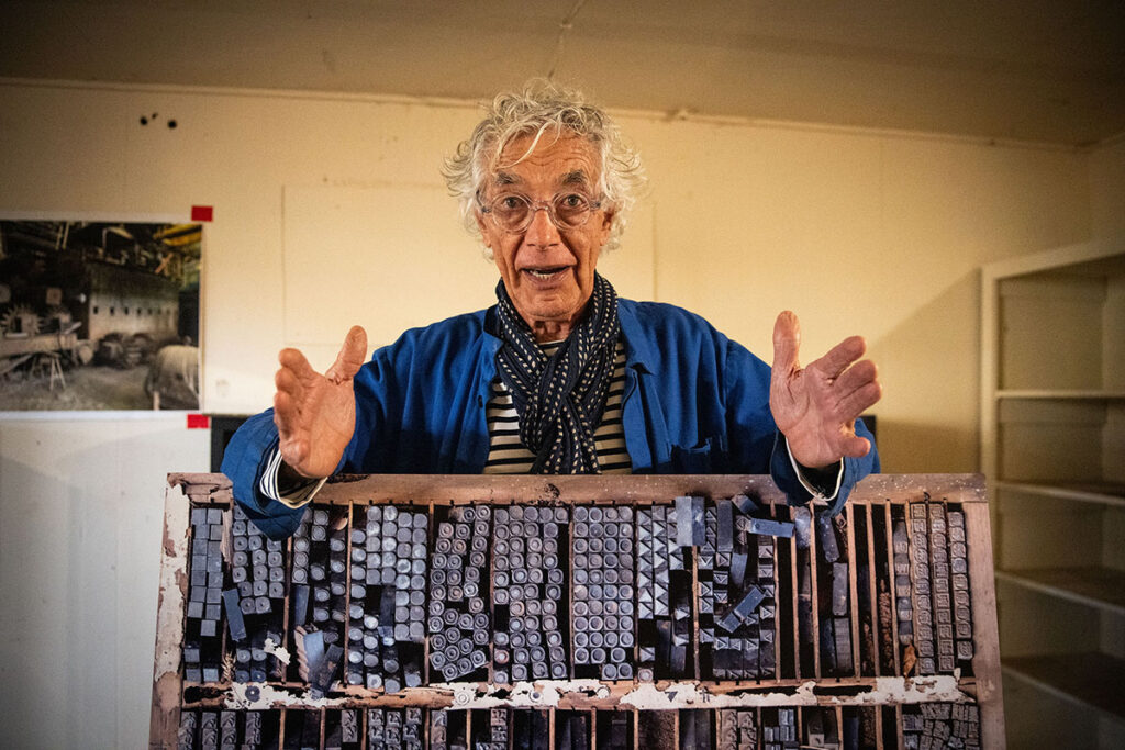 An elderly man with curly gray hair and glasses is gesturing while standing behind a tray filled with various types of metal letter stamps.