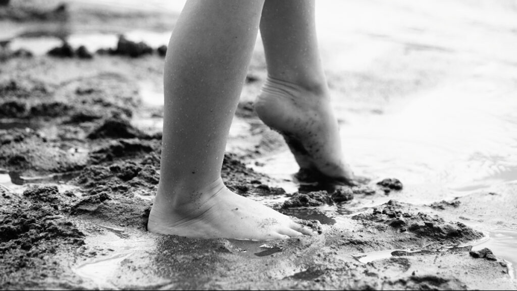 A close-up black and white photograph of a child’s bare feet as they walk through wet sand using cinematography lighting techniques to evoke a feeling of carefree exploration.