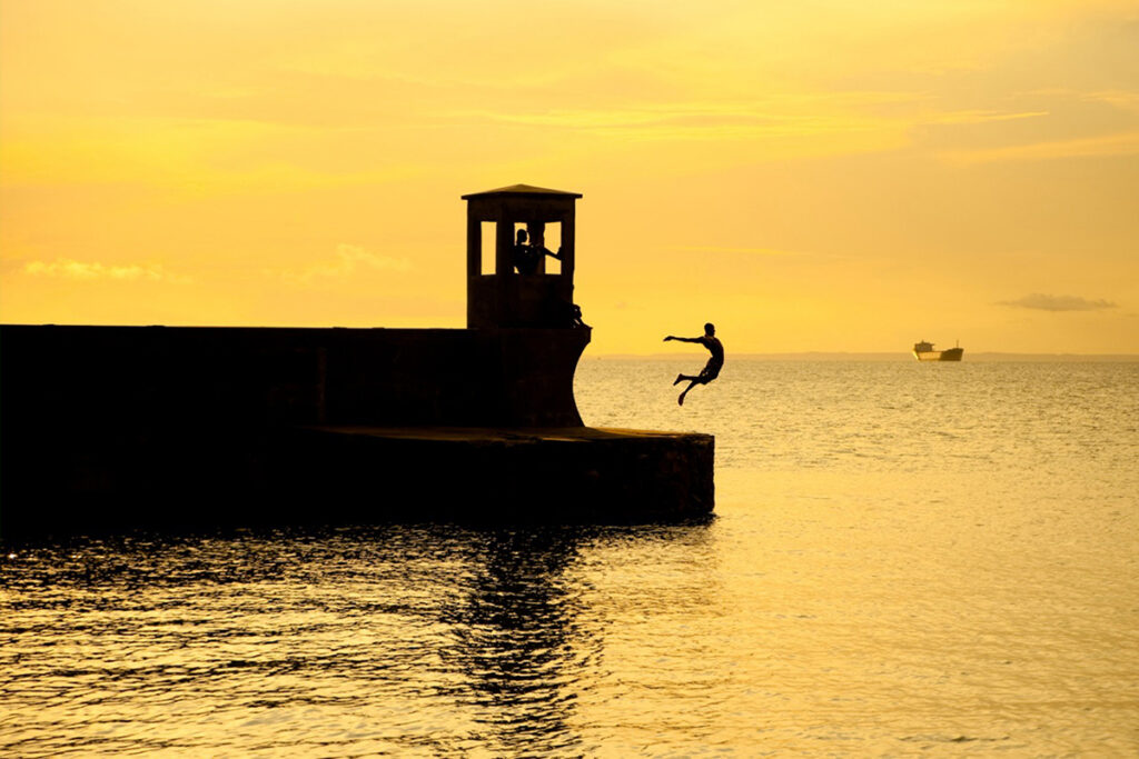 An example of cinematic videography showing a person in mid-air as they jump from a pier into the water during sunset.