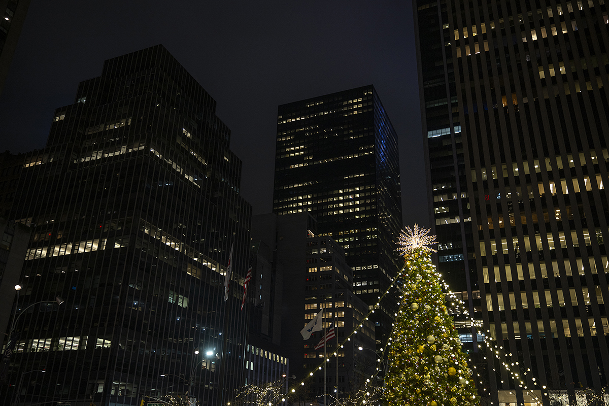 Uma árvore de Natal festiva adornada com luzes brilhantes se destaca contra o pano de fundo de prédios urbanos imponentes e iluminados.