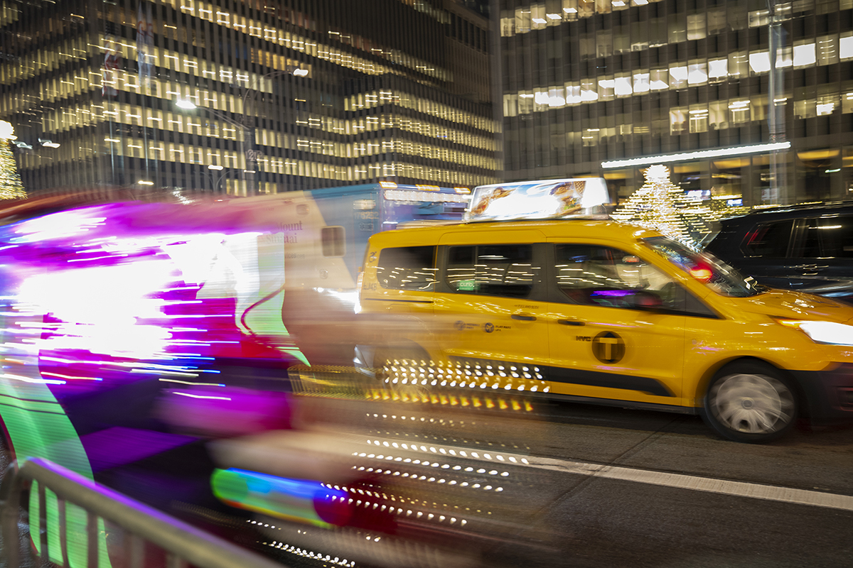 Um táxi amarelo clássico permanece parado enquanto luzes vibrantes e coloridas passam em um cenário urbano movimentado.