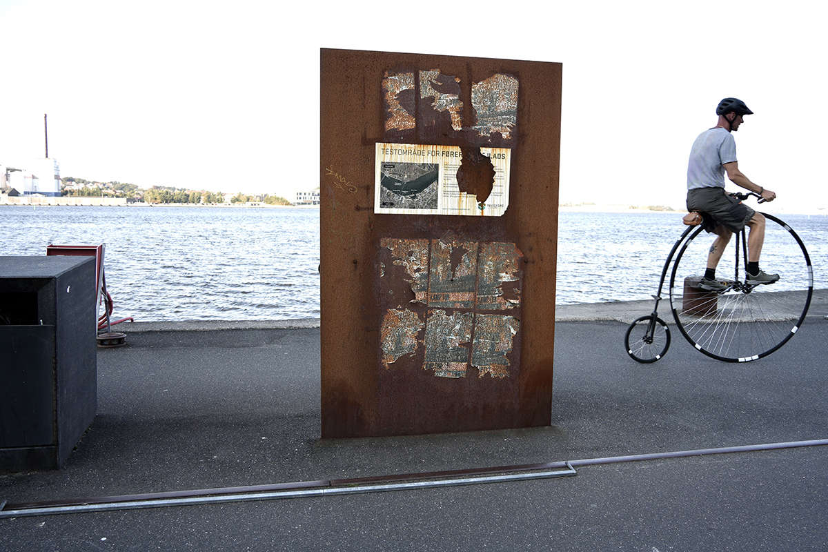 A man rides a vintage Penny-Farthing bicycle along the Aalborg waterfront in Denmark, passing a rusted metal sign with remnants of old posters.