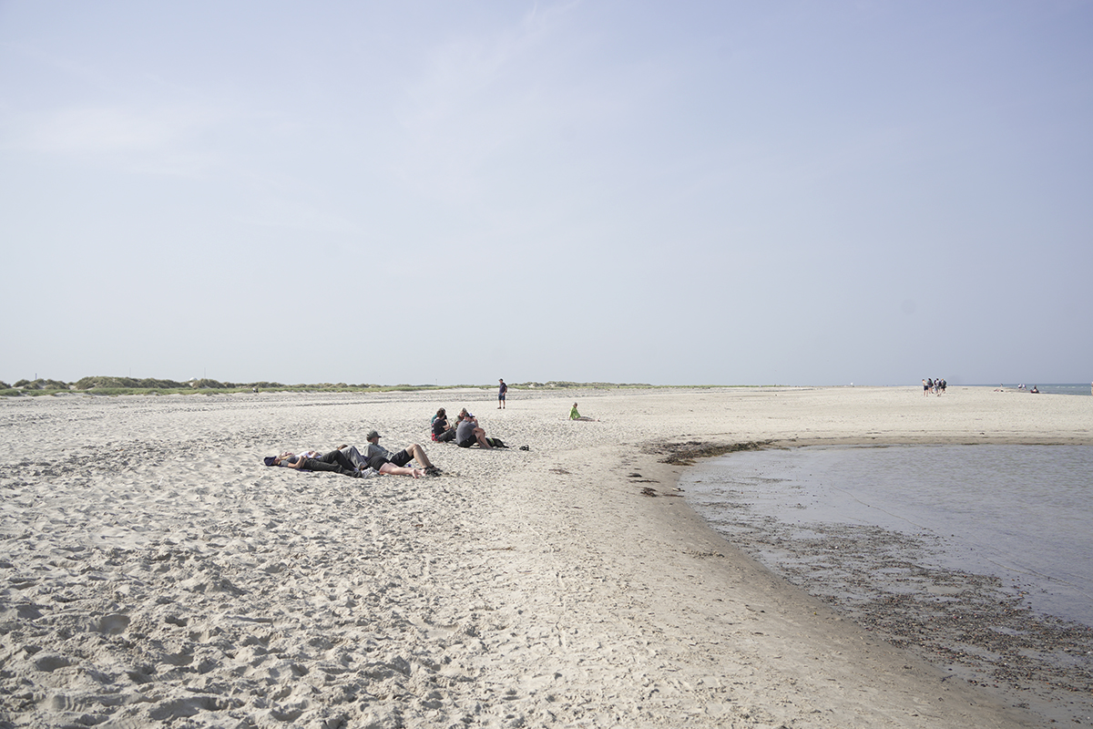 Des personnes se détendent et s'assoient le long d'une plage de sable près d'un bord d'eau peu profond, avec un ciel ouvert et des dunes lointaines en arrière-plan.