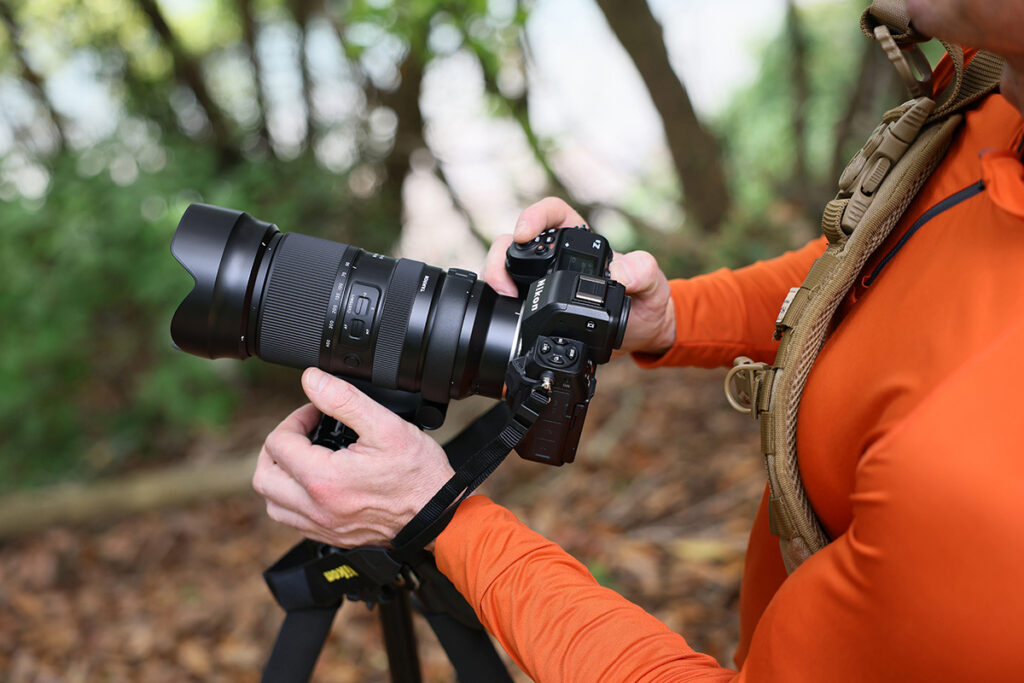 A photographer in an orange holding a camera mounted on a tripod in a natural forest setting. 
