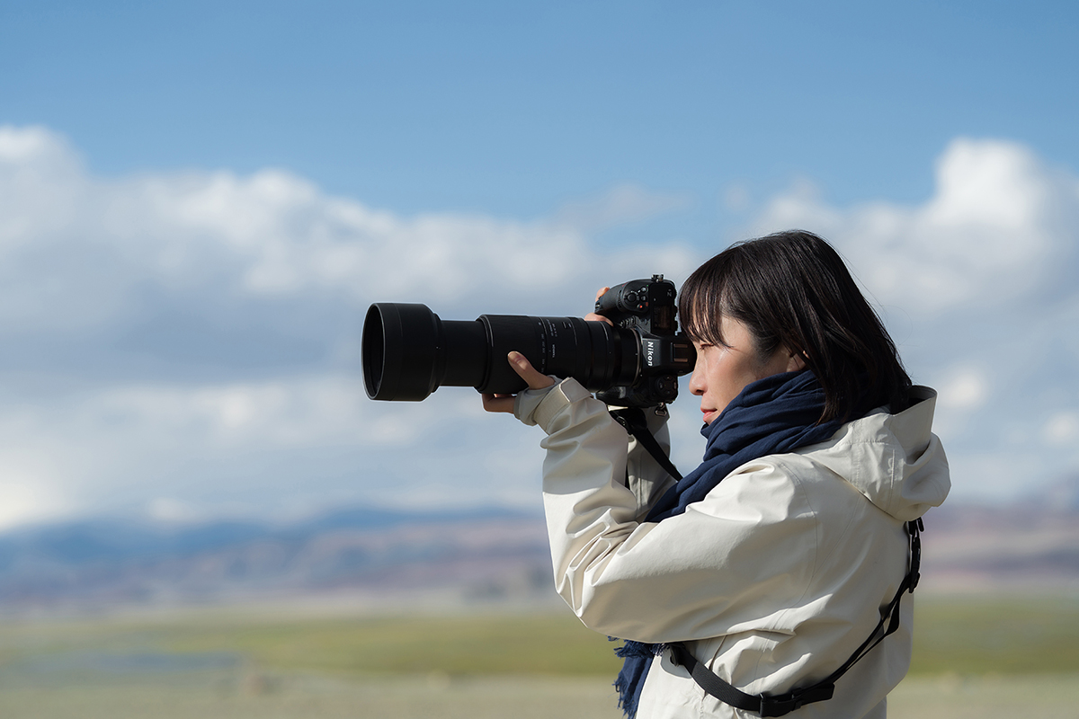 A photographer wearing a white jacket and blue scarf using a Nikon camera with a telephoto lens to capture a scenic outdoor view.