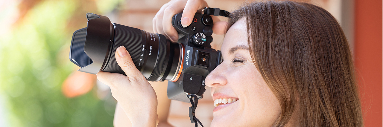 A smiling woman holds a camera with a Tamron lens, capturing a photograph outdoors.