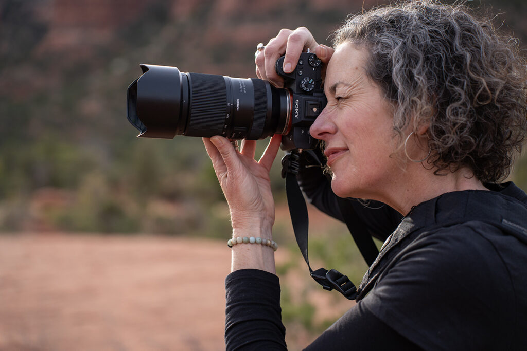 A woman using macro photography lens tips holds a camera to her eye to photograph a scenic outdoor landscape.