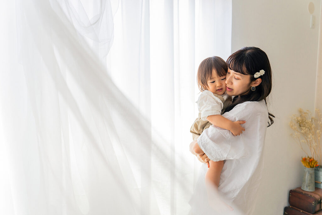 A woman holding a small child in a softly lit room with sheer white curtains.