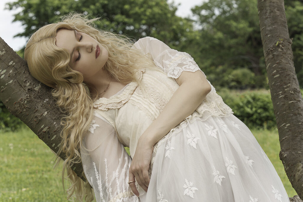 A serene image of a young woman wearing a flowing white dress resting against a tree trunk in a lush green outdoor setting.