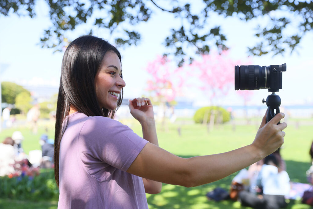 A smiling young woman records herself with a camera with one of the best lenses for Sony E-mount mounted on a handheld tripod in a sunny park.