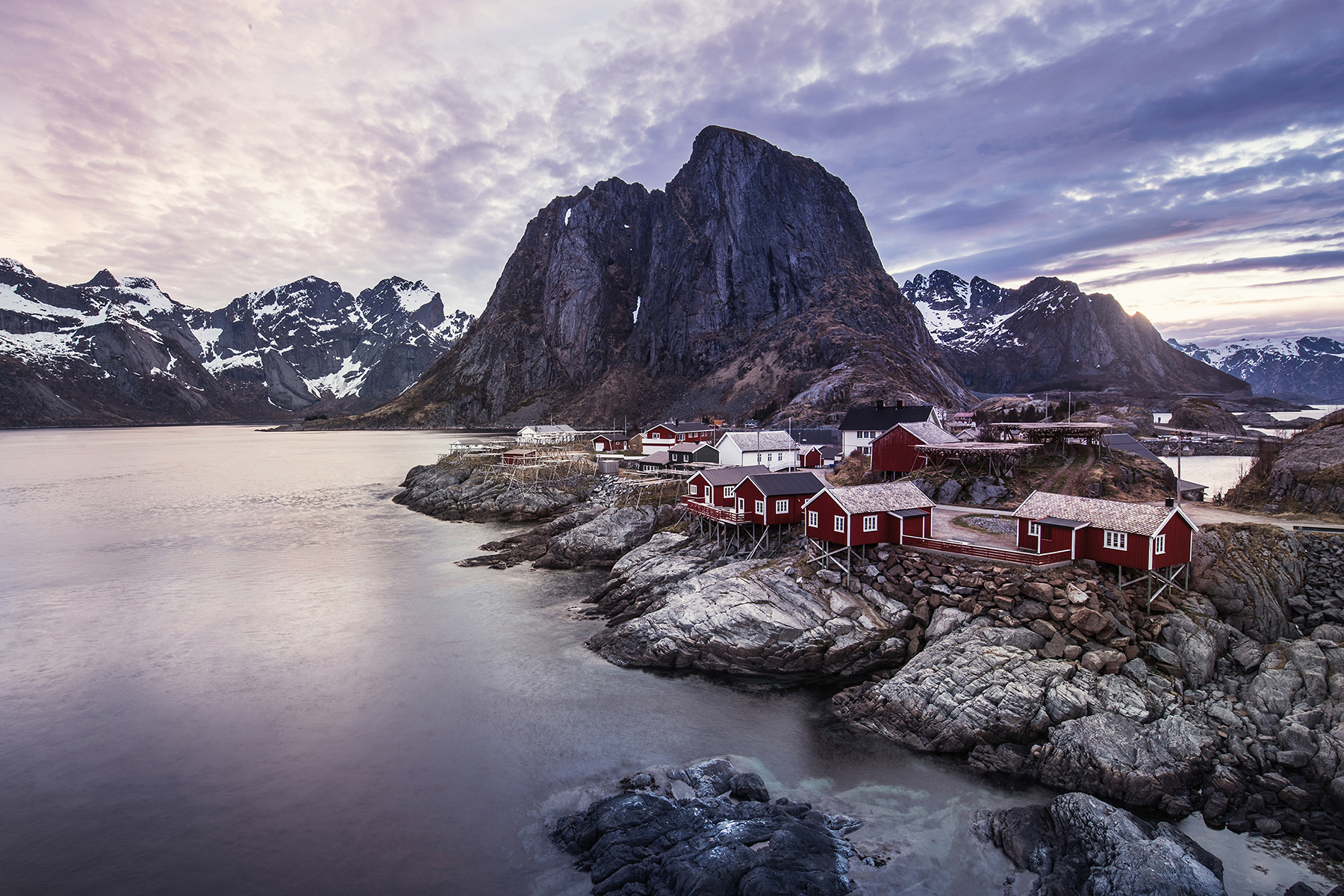 Idyllic scene of Lofoten's iconic red cabins nestled against dramatic cliffs and calm waters, under a serene twilight sky, captured with Tamron 24-70mm for DSLR cameras.