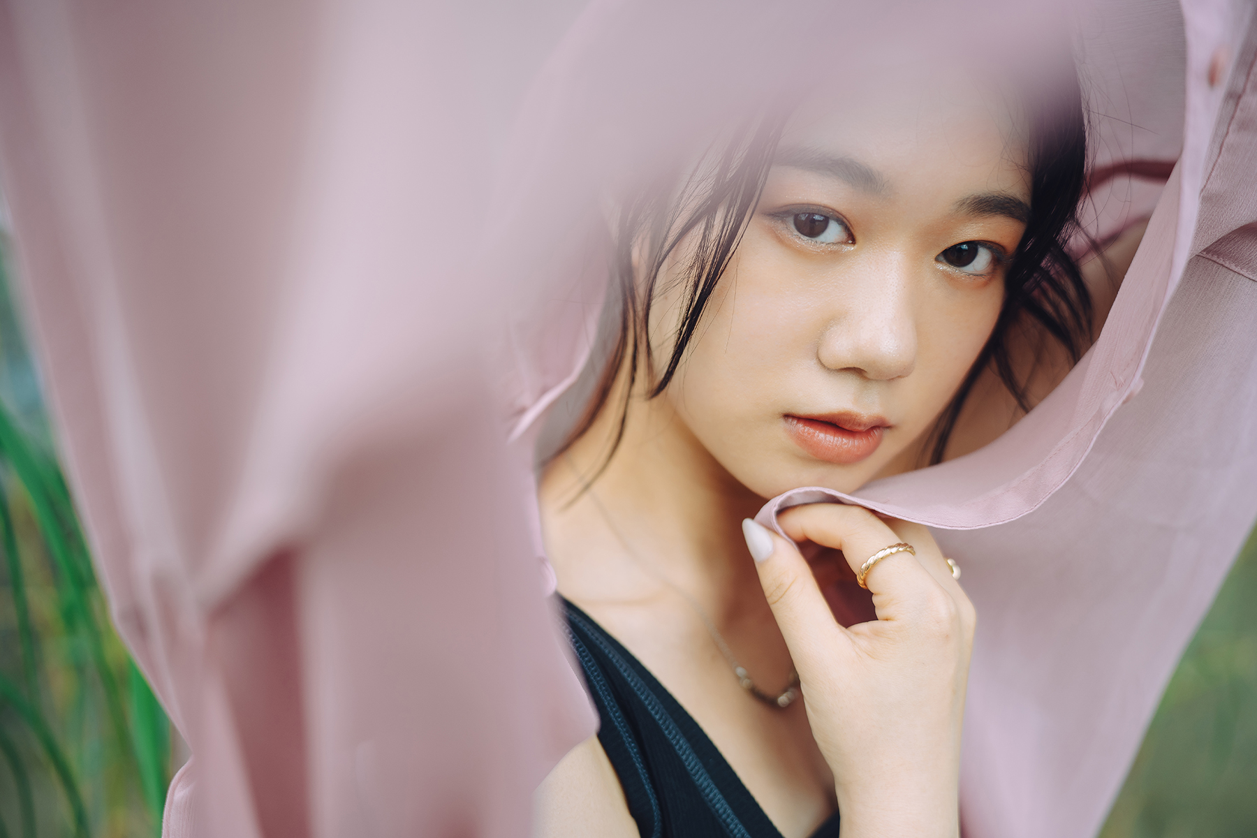 A close-up portrait of a young woman gazing softly at the camera, partially framed by flowing pink fabric.