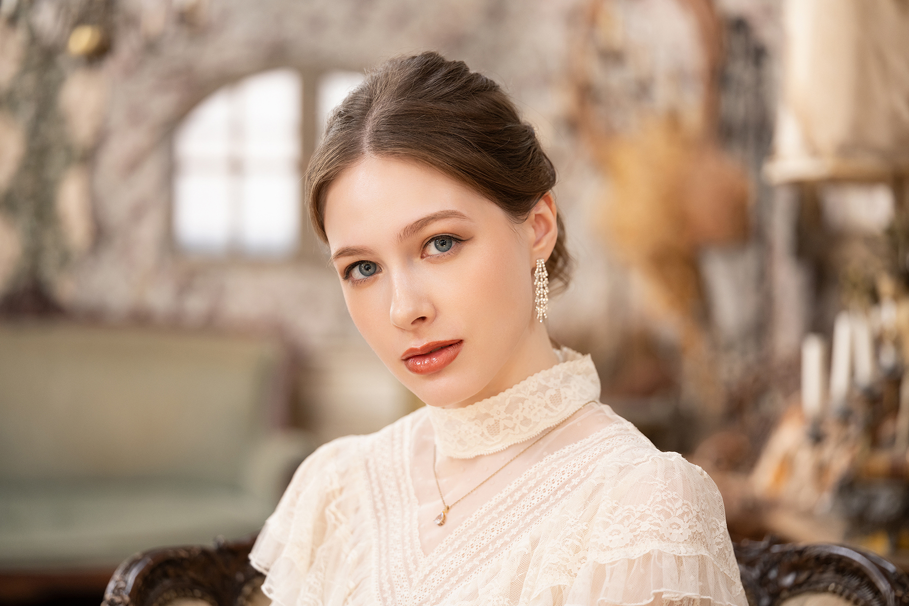 A portrait of a young woman in an elegant lace dress, seated in a vintage-style room with soft lighting and a blurred antique background.
