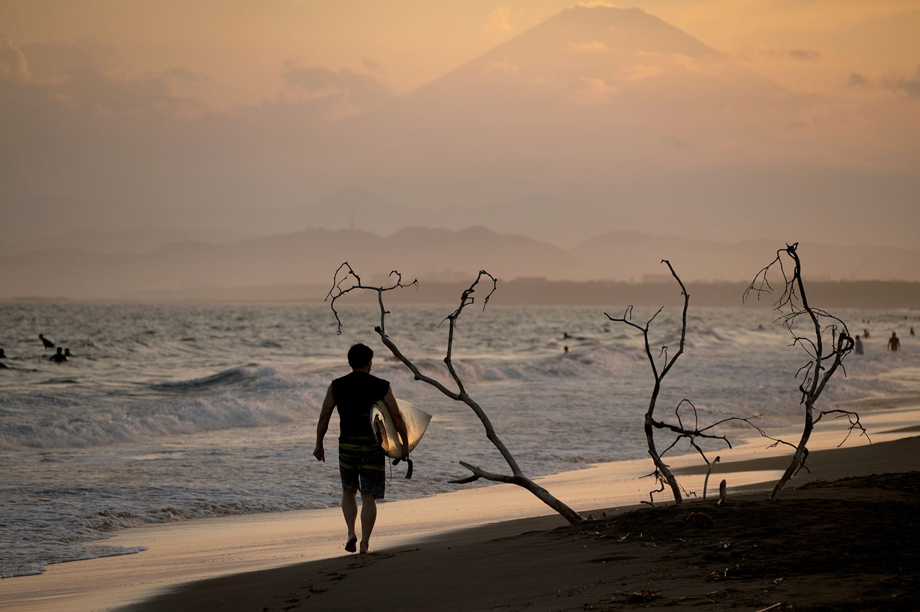 Un surfeur marche le long du rivage au coucher du soleil, portant une planche de surf reflétant la lumière dorée, avec des montagnes lointaines et l'océan en arrière-plan.