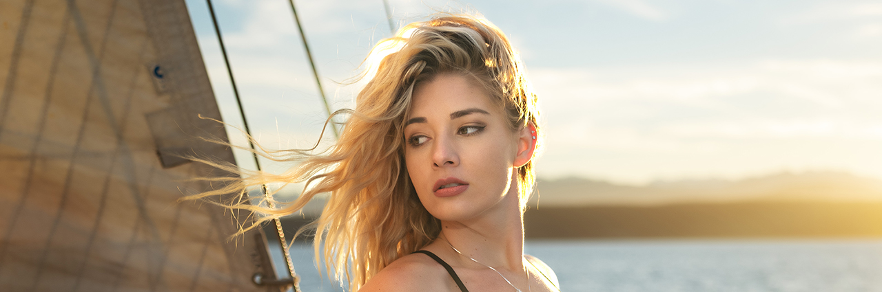 A young woman with windblown hair gazes into the distance aboard a sailboat at sunset.