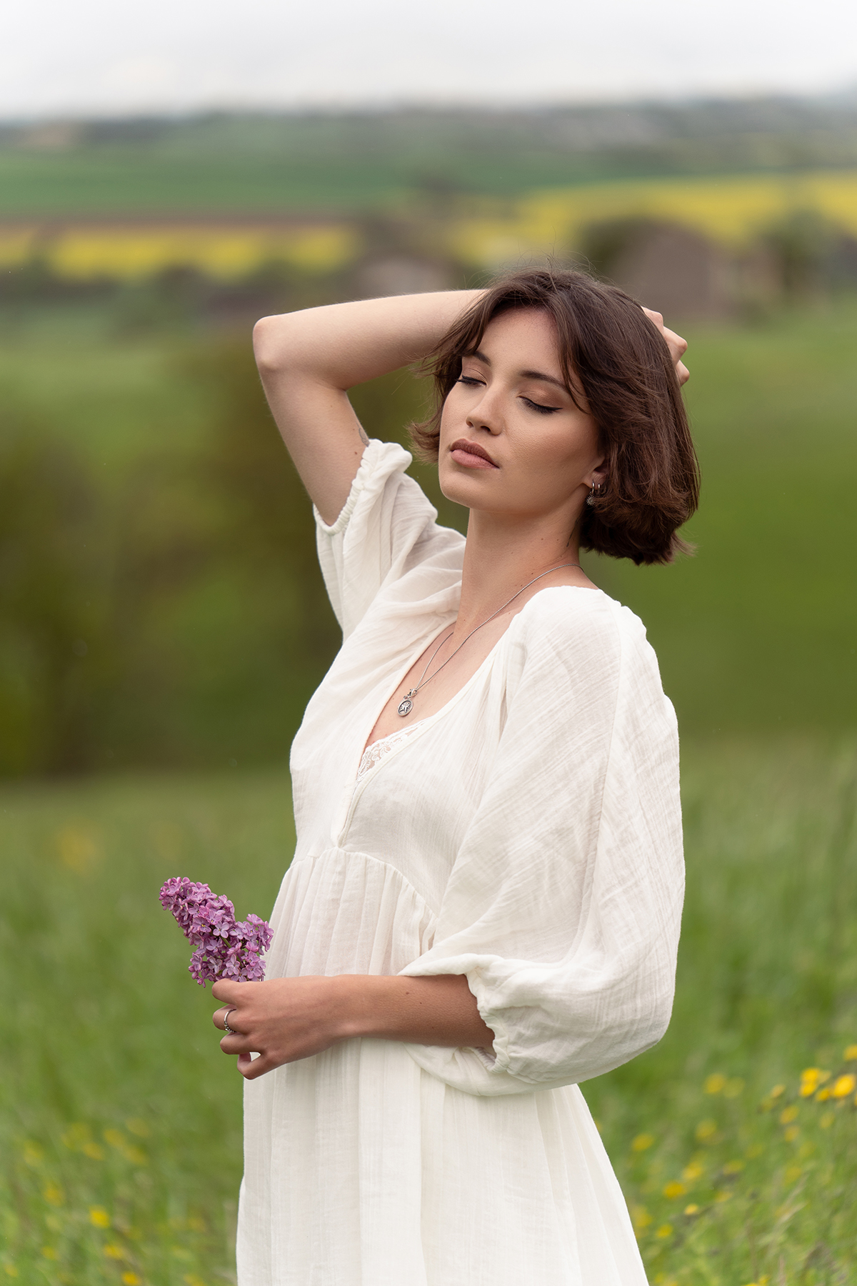 A young woman in a flowing white dress holding purple flowers, standing in a lush green field with a serene expression.