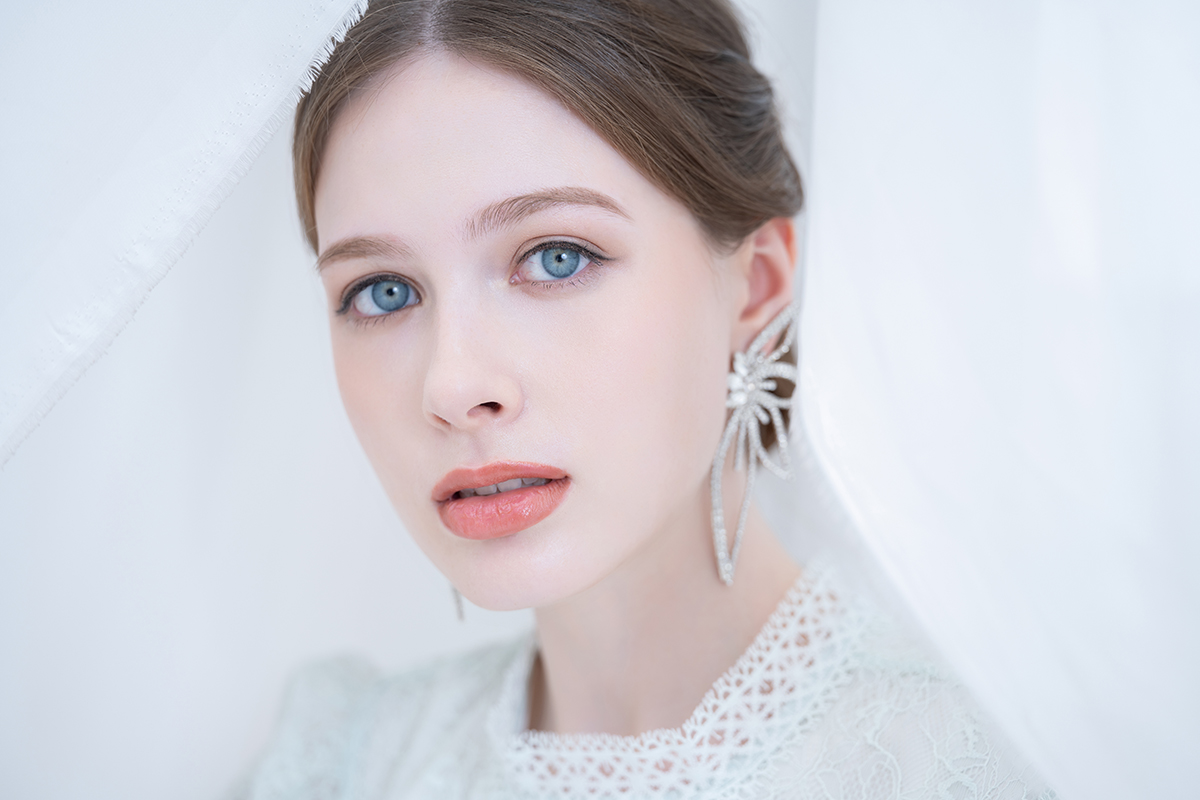 A young woman with blue eyes and elegant jewelry, framed by soft white fabric in a delicate, ethereal portrait.