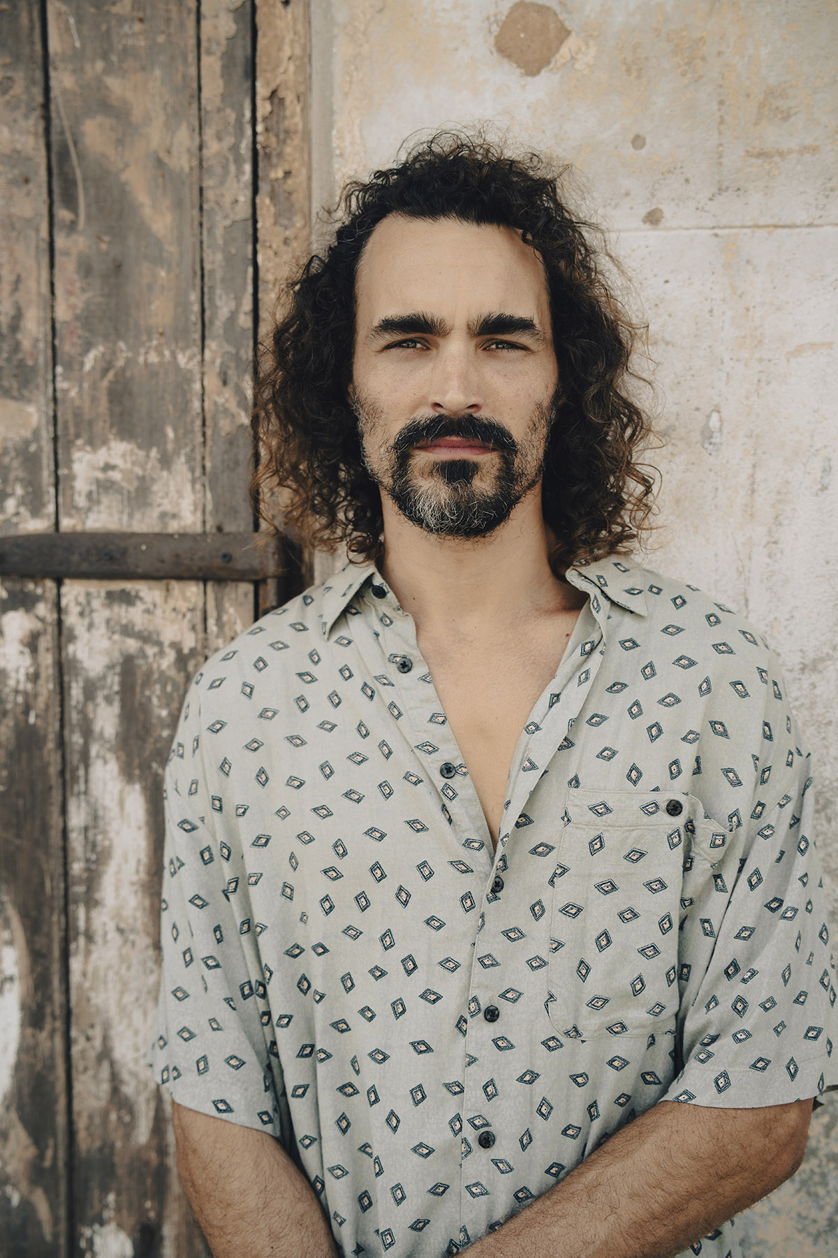 Portrait of a man with wavy hair and a patterned shirt standing against a rustic wooden and stone background, showcasing the depth and character captured with a high-quality travel portrait lens.