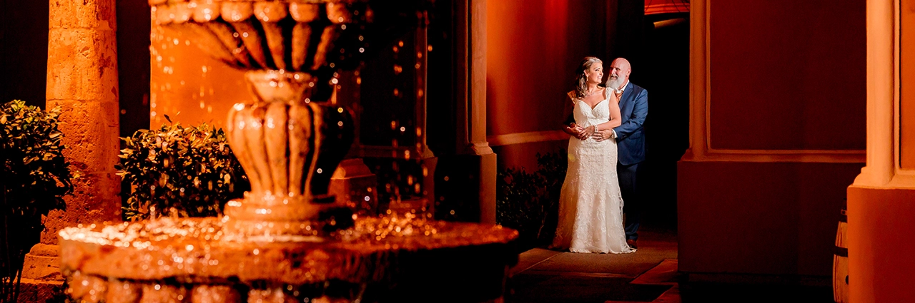 A newlywed couple poses elegantly in warm, dramatic lighting beside a cascading fountain, embodying the editorial wedding photography style with carefully curated composition and artistic direction.