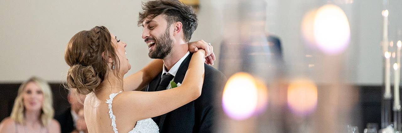 Bride and groom share a joyful first dance, captured in documentary style wedding photography with soft natural lighting and a blurred candle-lit foreground.