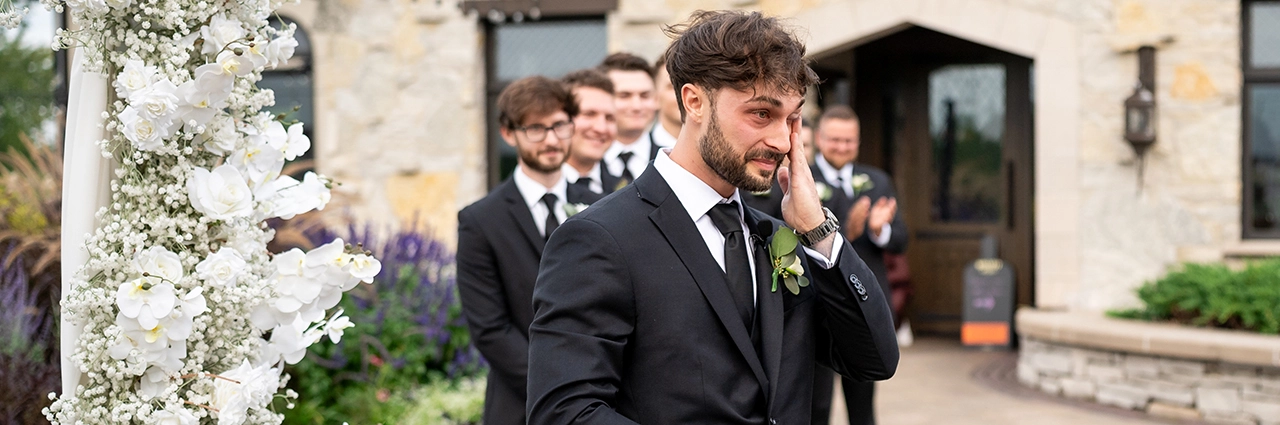 Groom overcome with emotion as he sees his bride walking down the aisle, captured in a candid documentary-style wedding moment with groomsmen cheering behind him.