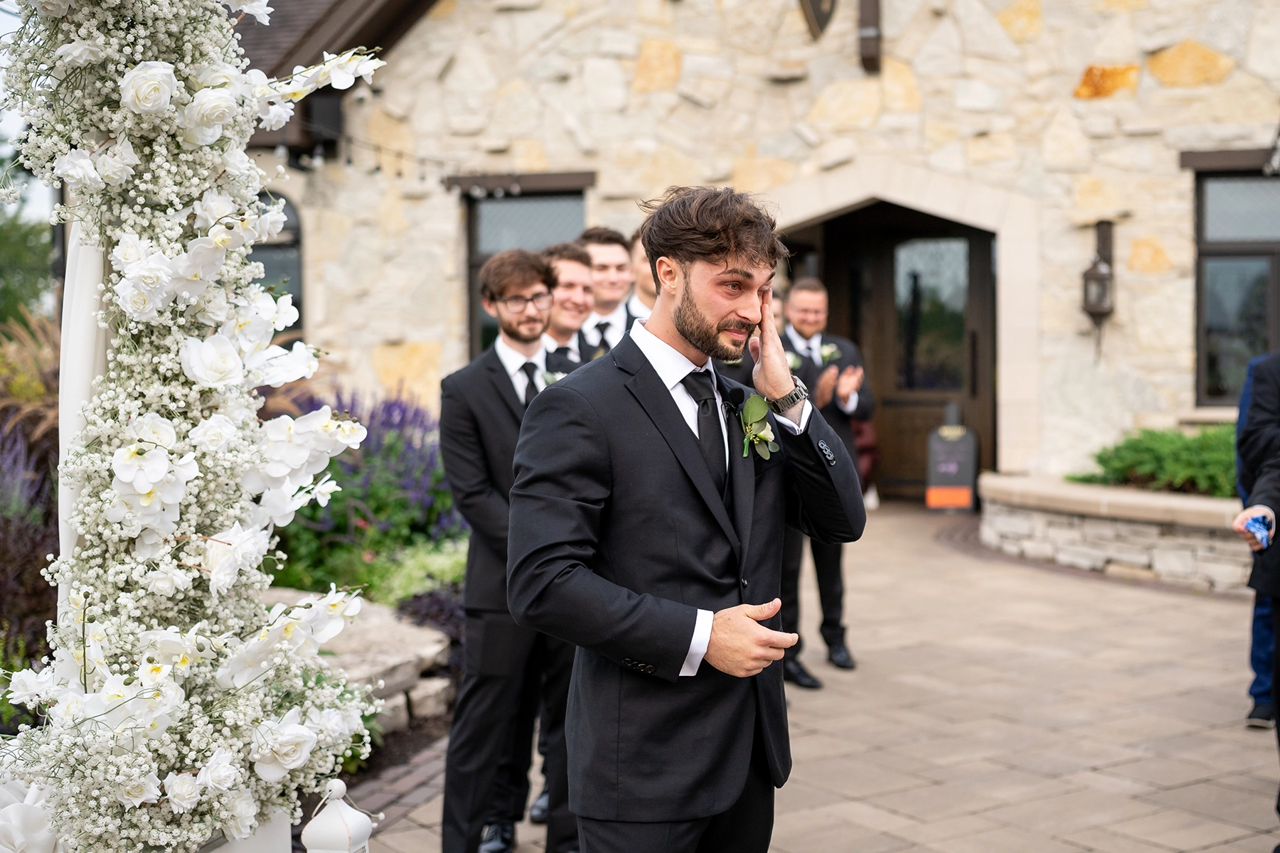 Emotional groom wiping away tears as he sees his bride for the first time during a documentary-style wedding ceremony, surrounded by groomsmen and floral decor.