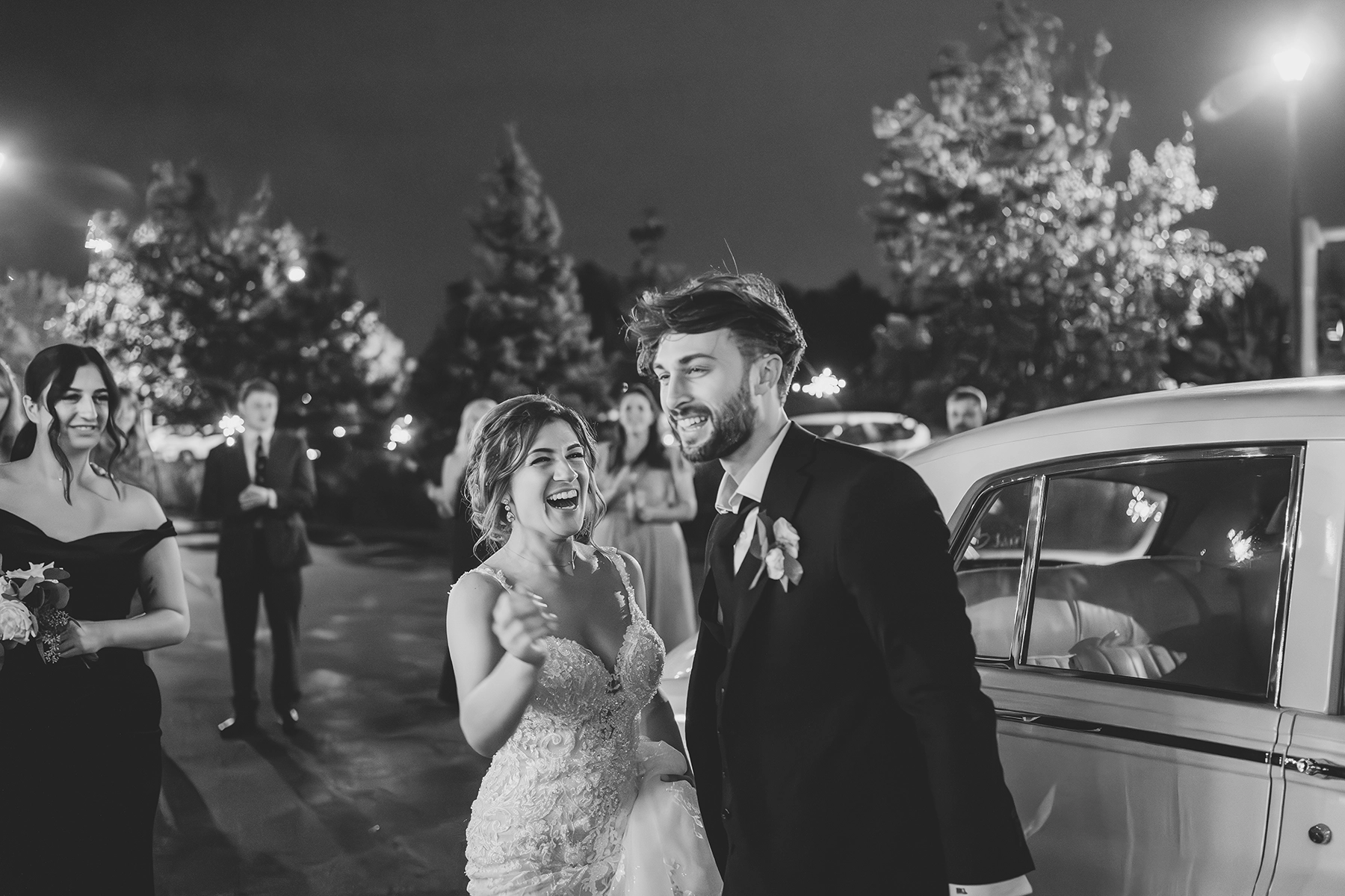 Black and white documentary-style wedding photography capturing a joyful moment as the bride and groom exit their reception, surrounded by loved ones.