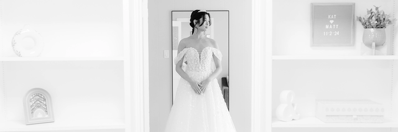 A bride stands gracefully in front of a mirror, her off-the-shoulder gown adorned with delicate floral details, as she is framed by softly lit white shelves—capturing the 2025 wedding photography trend of intimate, fashion-inspired bridal portraits.