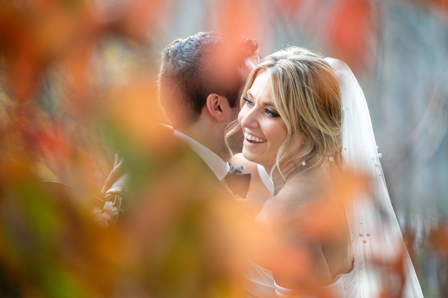 A bride and groom share an intimate moment, captured through autumn foliage. The bride, glowing with happiness, embraces her partner, reflecting 2025 wedding photography trends with a blend of documentary storytelling and soft editorial elegance.